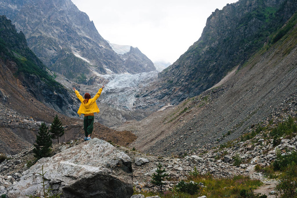 Reizen langs bergen, vrijheid en actieve levensstijl, bestemmingsconcept. Happy Woman toerist in geel jasje staand op berg in Svaneti, Georgia nabij Chalaadi gletsjer wandelroute - Foto, afbeelding