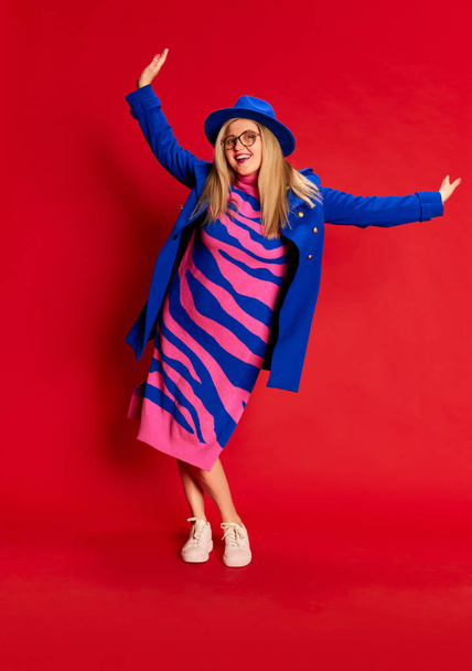 Happiness. Full-length portrait of woman in colorful stylish clothes emotionally posing, dancing against red studio background. Concept of emotions, facial expression, lifestyle and fashion - Foto, afbeelding
