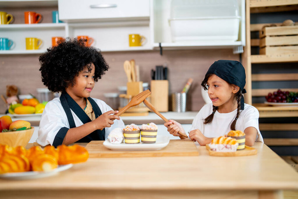 Zwei kleine Mädchen, afroamerikanische und asiatische Mädchen, benutzen Kochlöffel und Gabel, um während des Kochkurses in der Küche zu spielen und Spaß zu haben. - Foto, Bild