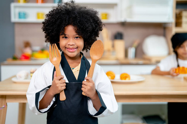 Мила афроамериканська дівчина тримає дерев "яну ложку і виделку і стоїть перед столом на кухні під час уроків приготування їжі.. - Фото, зображення