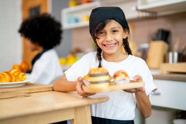 Little lovely girl hold plate contain cupcake and look at camera with smiling during study cooking class with her friend in kitchen. - Fotoğraf, Görsel