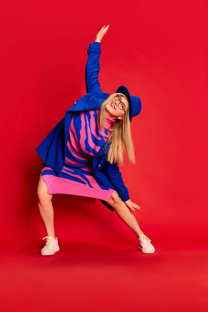 Happiness and positivity. Full-length portrait of woman in colorful stylish clothes emotionally posing, dancing against red studio background. Concept of emotions, facial expression, lifestyle and - Foto, afbeelding