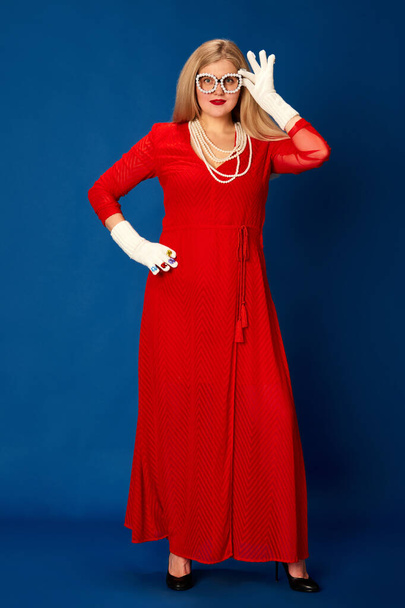 Portrait of beautiful woman in stylish red dress posing against blue studio background. Party and celebration. Concept of emotions, facial expression, lifestyle and fashion - Photo, image