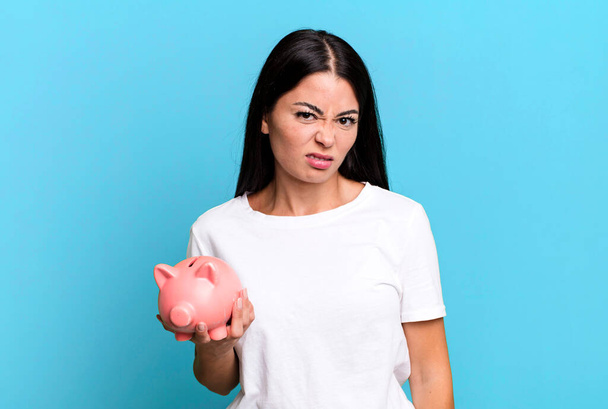hispanic pretty woman feeling puzzled and confused with a piggy bank - Fotoğraf, Görsel