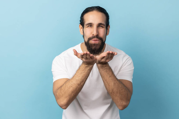 Portrait of man with beard wearing white T-shirt blowing air kiss, folded palm towards mouth, sends mwah coquettish, wants to cheer up his partner. Indoor studio shot isolated on blue background. - Fotó, kép