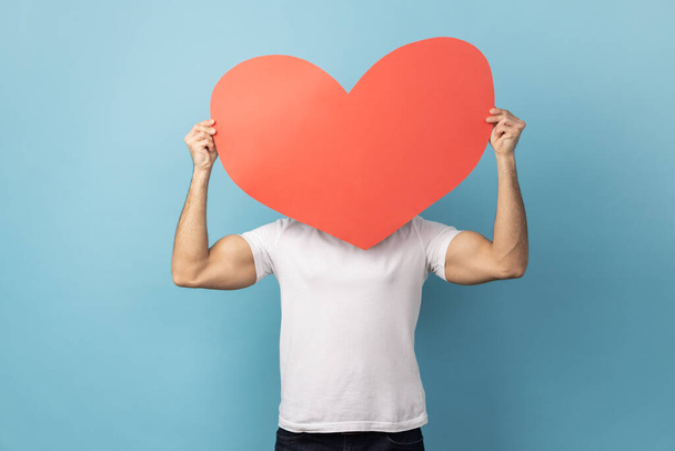 Portrait of man wearing white T-shirt hiding behind big red paper heart, holding symbol of love care, cardiac issues, demonstrating affection. Indoor studio shot isolated on blue background - Fotó, kép