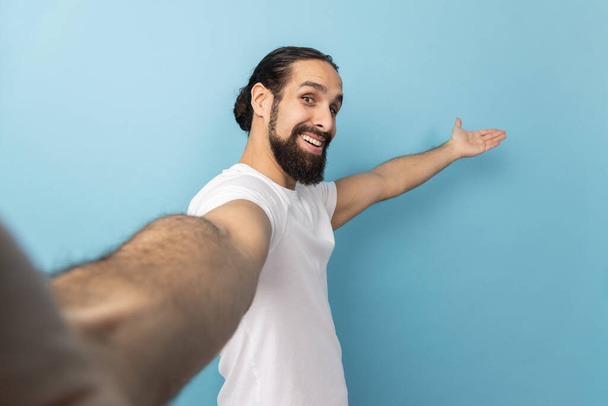 Portrait of king friendly man with beard wearing white T-shirt looking at camera with smile, making welcome gesture, taking selfie POV. Indoor studio shot isolated on blue background. - Фото, зображення