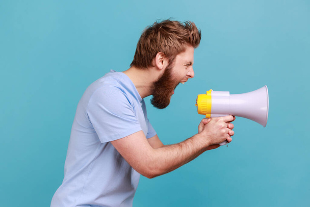 Side view of bearded man holding megaphone near mouth, loudly speaking, screaming, making announcement, paying attention at social problems. Indoor studio shot isolated on blue background. - Photo, image