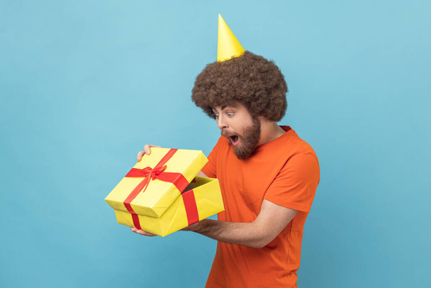 Portrait of surprised man with Afro hairstyle wearing orange T-shirt and party cone unpacked present box, looking inside with open mouth. Indoor studio shot isolated on blue background. - Valokuva, kuva