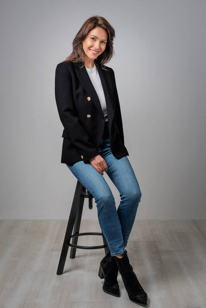 Full length studio shot of a beautiful smiling woman sitting at isolated grey background. Happy woman wearing black blazer with white shirt and blue jeans. Copy space. - Foto, Imagem