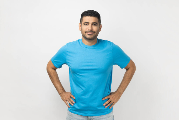Portrait of happy delighted optimistic unshaven man wearing blue T- shirt standing looking at camera, expressing positive emotions, keeps hands on hips. Indoor studio shot isolated on gray background. - Foto, Imagen