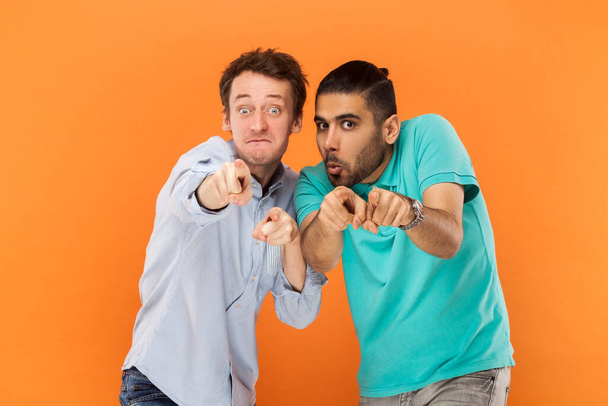 Portrait of two comic amazed men friends standing and pointing at camera, choosing you, having finny surprised expressions. Indoor studio shot isolated on orange background. - Fotoğraf, Görsel