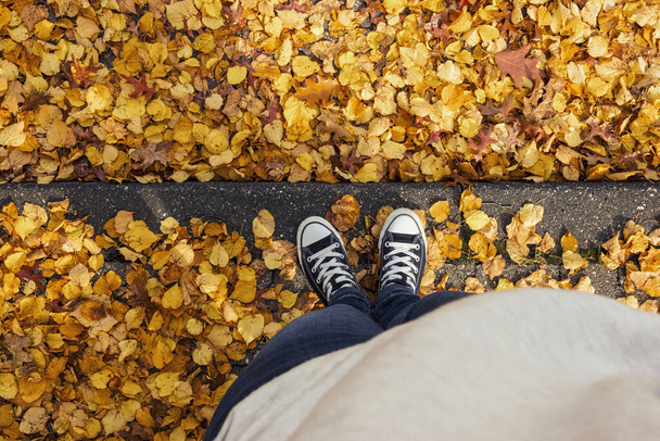 Young woman in sneakers stands in at a curbstone with foliage leafs at autumn on the streets, personal pespective from above. - Photo, Image