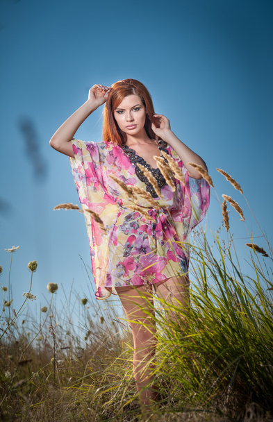 Beautiful young woman in wild flowers field on blue sky background. Portrait of attractive red hair girl with long hair relaxing in nature, outdoor shot. Lady in multicolored dress enjoying nature - Photo, Image