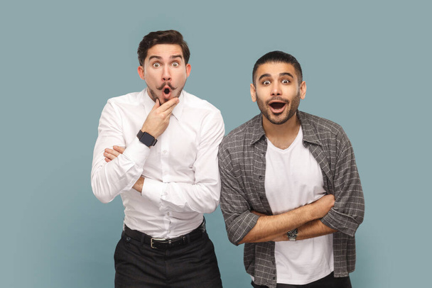 Portrait of two shocked men friends standing together, see something astonished, keeping mouth opened and looking with big eyes. Indoor studio shot isolated on light blue background. - Foto, Imagen