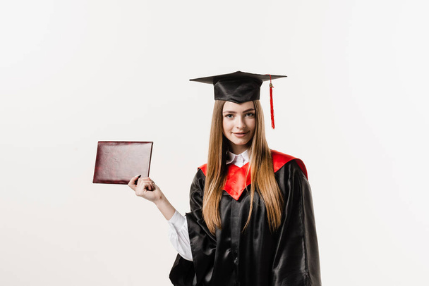 Graduate girl with master degree diploma in black graduation gown and cap on white background. Happy young woman successfully graduated from the university with honors - Photo, Image