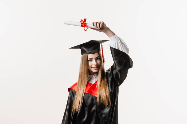 Graduate girl is graduating college and celebrating academic achievement. Happy girl student in black graduation gown and cap raises masters degree diploma above head on white background - Foto, immagini