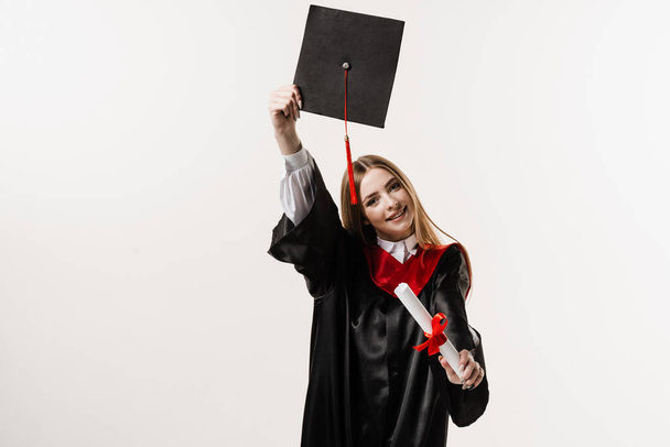 Graduate girl is graduating high school and celebrating academic achievement. Masters degree diploma in hands. Happy student in black graduation gown and cap is smiling on white background - Zdjęcie, obraz
