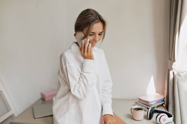 Charmante europäische Mädchen mit hellen Haaren tragen weißes Hemd sitzt auf dem Tisch im weißen Raum und telefoniert mit lieblichem Lächeln  - Foto, Bild