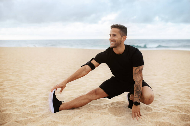 Молодой сильный спортсмен в спортивной одежде занимается разминкой, разминает ноги на пляже у моря на открытом воздухе, смотрит в сторону и улыбается, свободное место - Фото, изображение