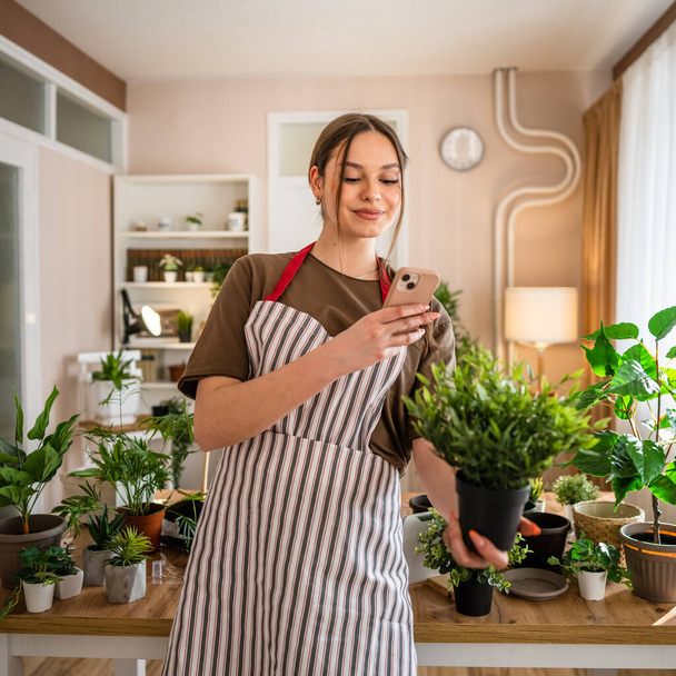 Uma mulher jovem geração z adulto caucasiano feminino cuidar de suas plantas em casa fotografar vaso de flores com seu telefone celular smartphone enviar fotos para as mídias sociais ou como pessoa real mensagem - Foto, Imagem