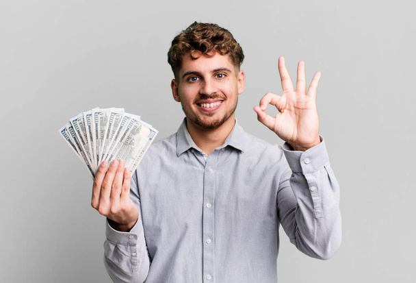 young adult caucasian man feeling happy, showing approval with okay gesture. dollar bank notes concept - Zdjęcie, obraz
