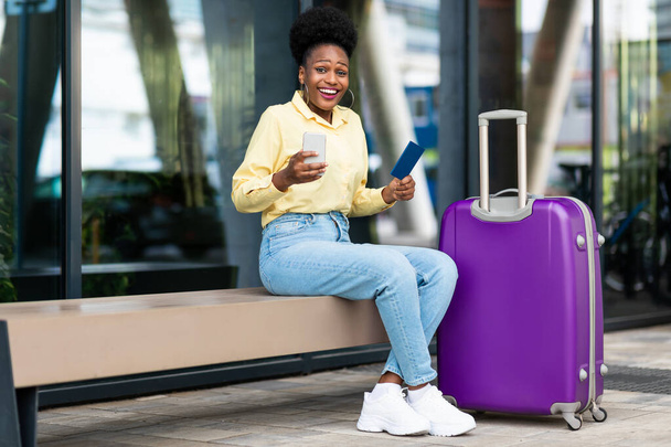 Aufgeregte afroamerikanische Touristin, die per Handy Reisetickets bucht und in die Kamera lächelt, während sie mit dem Koffer im Freien in der Nähe des Flughafenterminals sitzt. Endlich Urlaub - Foto, Bild
