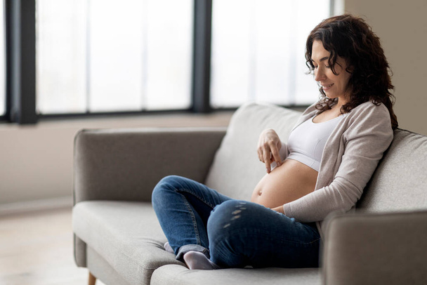 Portrait of young pregnant woman walking with fingers acroos her big belly while sitting on couch at home, smiling expectant female preparing for maternity, enjoying her pregnancy time, copy space - Foto, Bild
