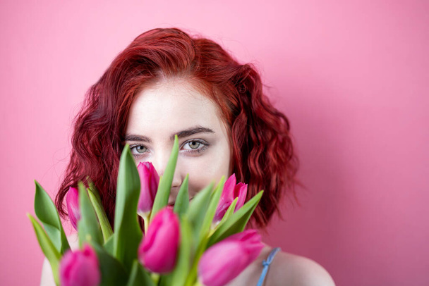 Portré egy gyönyörű fiatal vörös hajú lány kezében egy csokor íriszek és tulipánok elszigetelt rózsaszín háttér - Fotó, kép