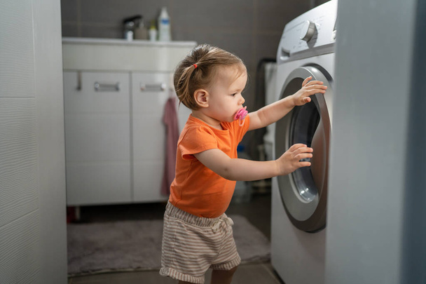 One girl small caucasian toddler child daughter standing at the washing machine in the toilet opening or closing the door examine and learn early development and growing up mischief concept copy space - Photo, image