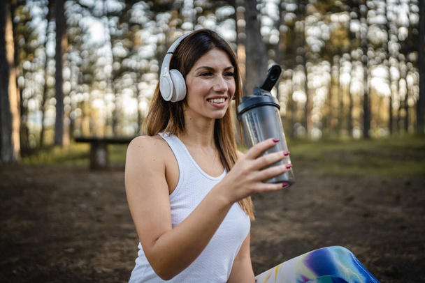 One woman happy caucasian female sitting in the forest or park in woods with supplement shaker and headphones during training in nature taking a brake water drink and hydration real people copy space - Fotó, kép