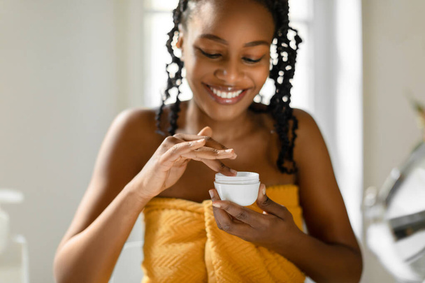 Daily skincare rotine. Happy black woman holding cream jar and applying moisturizer, selective focus on hands. Lady taking care of aging skin - Foto, Bild