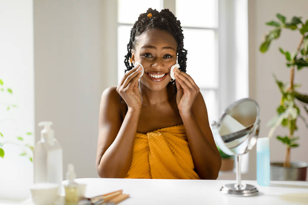 Onnellinen kaunis musta nainen istuu peilin edessä makuuhuoneessa, pyyhkimällä meikkiä kasvoiltaan, käyttämällä puuvillatyynyjä ja puhdistusvoidetta, hymyilemällä kameralle - Valokuva, kuva