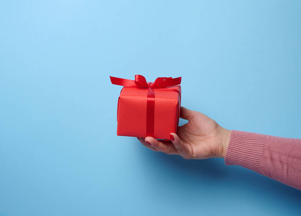 Une main de femme tenant une boîte cadeau rouge enveloppée d'un ruban sur un fond bleu, un concept de félicitations, surprise - Photo, image