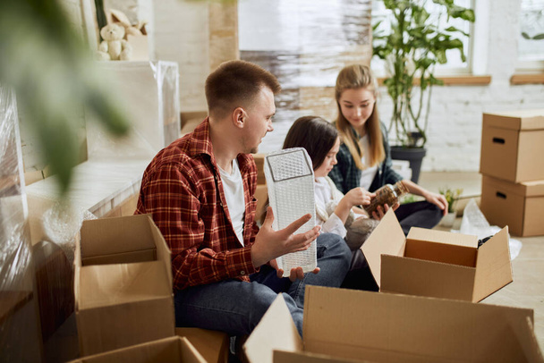 Young happy family, man, woman and kid moving into new flat, apartment. Opening many cardboard boxes with stuffs in living room on daytime. Concept of moving houses, real estate, family, new life - Photo, Image