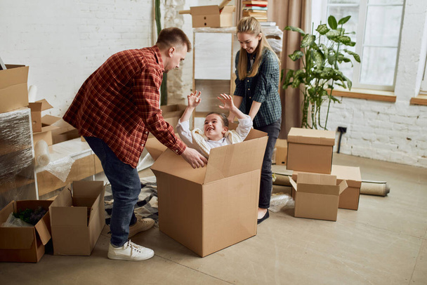 Young happy family, man, woman and kid moving into new flat, apartment with many cardboard boxes. Daughter hiding into box. Playing. Concept of moving houses, real estate, family, new life - Фото, зображення