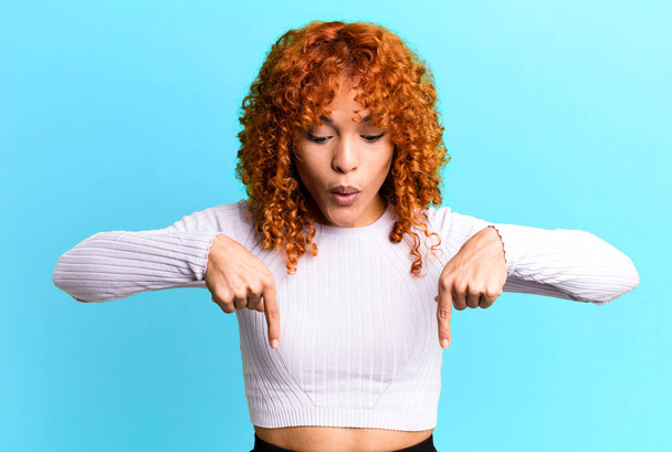 redhair pretty woman with open mouth pointing downwards with both hands, looking shocked, amazed and surprised - Foto, Bild
