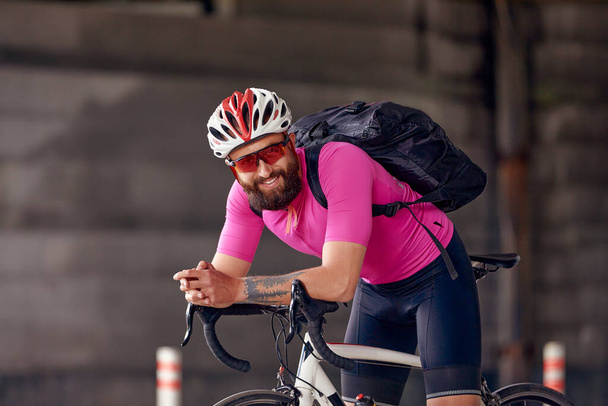 Um ciclista profissional em sportswear, um capacete de segurança e óculos está se preparando para um passeio de bicicleta matinal na arquitetura de concreto urbano. O conceito de um estilo de vida ativo e saudável - Foto, Imagem