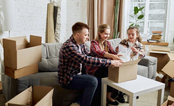 Putting things on place. Young happy family, man, woman and kid moving into new flat, apartment with many cardboard boxes. Concept of moving houses, real estate, family, new life - Photo, Image