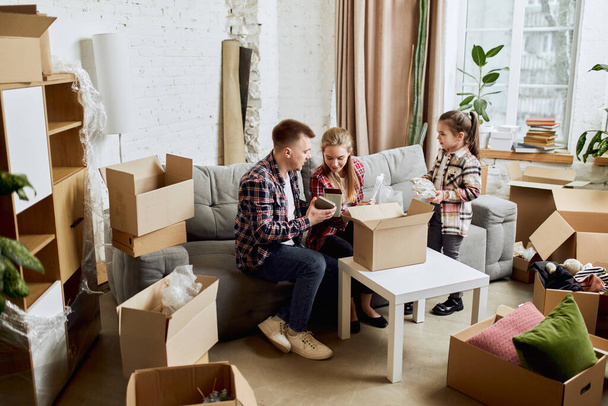 Putting things on place. Young happy family, man, woman and kid moving into new flat, apartment with many cardboard boxes. Concept of moving houses, real estate, family, new life - Foto, imagen