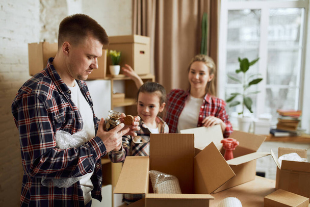 Young happy family, man, woman and kid moving into new flat, apartment with many cardboard boxes filled with stuffs and clothes. Concept of moving houses, real estate, family, new life - Photo, Image