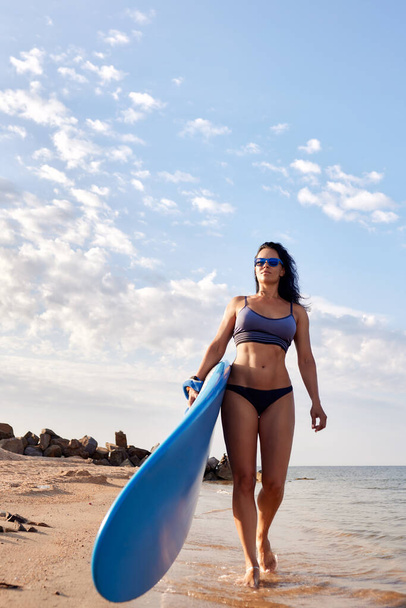 Charming woman with dark hair and athletic tanned body smiling carries a windsurfboard in the setting sun with a surfboard on the beach on a sunny summer day, ready to surf. in a bathing suit. - Foto, Imagem