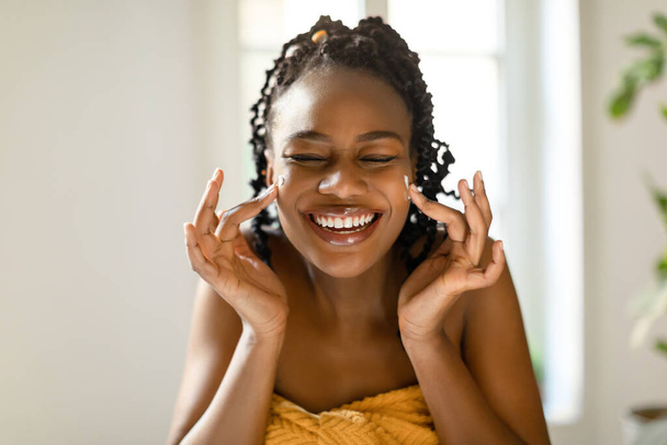 Όμορφη μαύρη γυναίκα που αγγίζει το πρόσωπό της και χαμογελά, κάνει θεραπείες ομορφιάς στο σπίτι, Αφροαμερικανή κυρία που κάθεται τυλιγμένη σε πετσέτα και απολαμβάνει την αυτοφροντίδα - Φωτογραφία, εικόνα