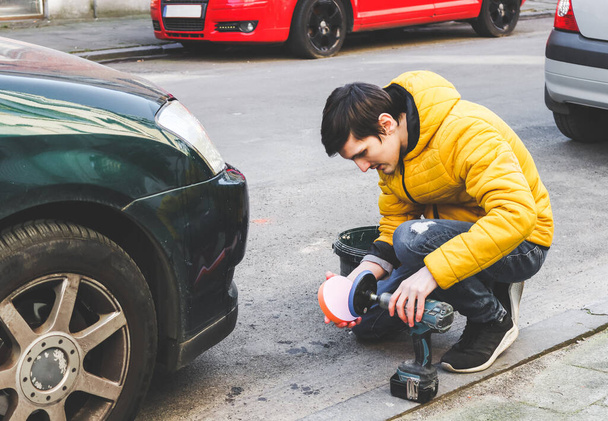 Ein junger Kaukasier in gelber Jacke und Jeans legt halb sitzend eine runde Schwammscheibe auf eine Bohrmaschine, um die Scheinwerfer seines Autos auf einer Stadtstraße vor dem Haus zu polieren, Nahaufnahme von der Seite. Das Konzept der Autowäsche zu Hause, Scheinwerferreinigung,  - Foto, Bild