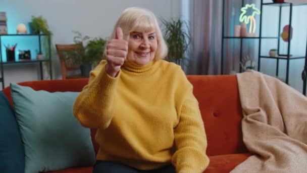 Mint például. Boldog izgatott idős nő, aki helyeslően néz a kamerába, hüvelykujját felfelé mutatva, mint valami pozitív jel, jó hír, pozitív visszajelzés. Az idősebb nagymama otthon ül a kanapén. - Felvétel, videó