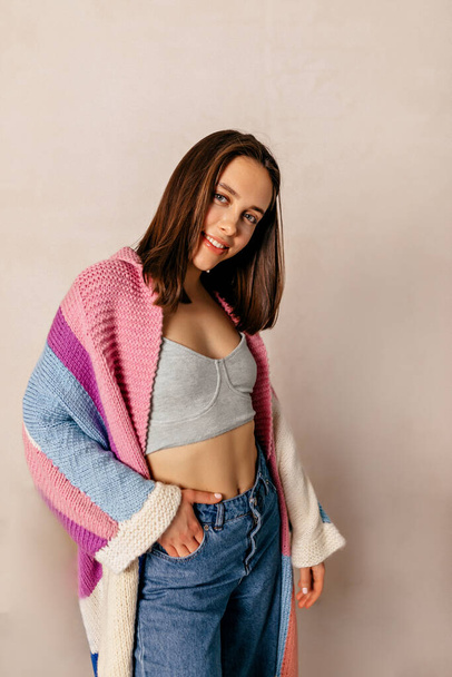 Happy young stylish girl in good mood posing at camera with putting arms in pockets on beige background. Brunette woman with short hair in bright knitted pullover. Weekend enjoyment concept  - Foto, afbeelding