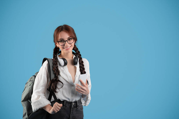 Blij tiener meisje student met staarten en rugzak in bril en koptelefoon typen op smartphone op blauwe studio achtergrond. App voor studie en kennis op school, advertentie en aanbod, nieuw normaal - Foto, afbeelding