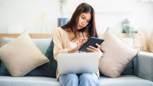 自宅のソファに座っている間、コンピュータのラップトップと携帯電話のスマートフォンを使用して若いアジアの女性 - 写真・画像