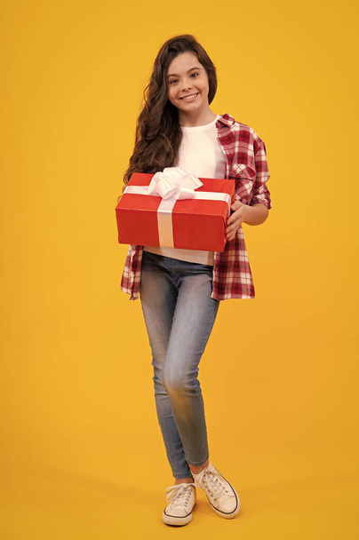 Happy teenager, positive and smiling emotions of child girl. Teenager kid with present box on isolated yellow background. Teen girl giving birthday gift. Present, greeting and gifting concept - Fotoğraf, Görsel