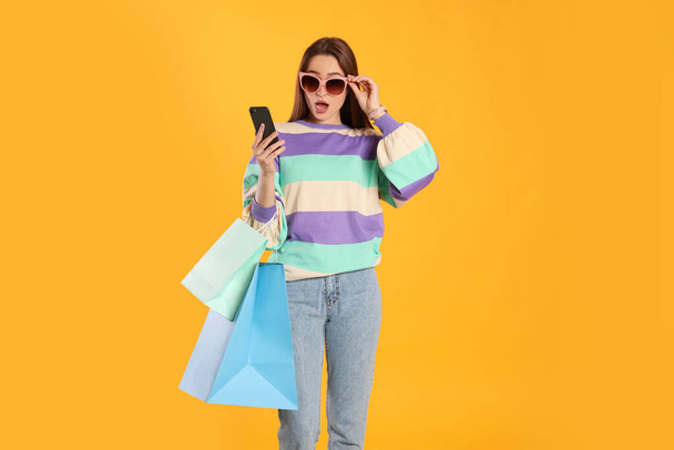 Surprised young woman with shopping bags looking at smartphone on yellow background. Big sale - Zdjęcie, obraz